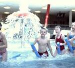 Fünf Kinder, die Poolnudeln halten, spielen in einem Hallenbad mit einem Wasserspiel im Hintergrund.