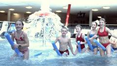 Fünf Kinder, die Poolnudeln halten, spielen in einem Hallenbad mit einem Wasserspiel im Hintergrund.