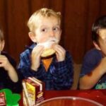 Drei Jungen sitzen an einem Tisch und essen jeweils einen gepuderten Donut mit weißem Puder im Mund. Auf dem Tisch vor ihnen stehen Kisten mit Schokoladenmilch.