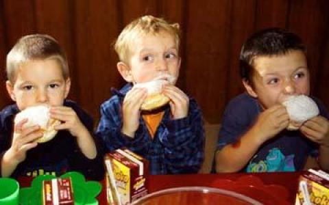 Drei Jungen sitzen an einem Tisch und essen jeweils einen gepuderten Donut mit weißem Puder im Mund. Auf dem Tisch vor ihnen stehen Kisten mit Schokoladenmilch.