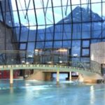Hallenbad mit geschwungener Brücke, roten Liegestühlen und großen Glasfenstern mit Blick auf die schneebedeckten Berge draußen.