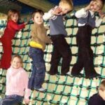 Sechs Kinder klettern im Haus an einer Seilnetzwand hoch, während ein Kind am Boden sitzt und alle zu spielen scheinen.