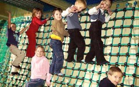 Sechs Kinder klettern im Haus an einer Seilnetzwand hoch, während ein Kind am Boden sitzt und alle zu spielen scheinen.