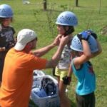 Ein Erwachsener hilft einer Gruppe von Kindern beim Anziehen von Helmen und Handschuhen auf einer Wiese, um sie auf eine Aktivität vorzubereiten.