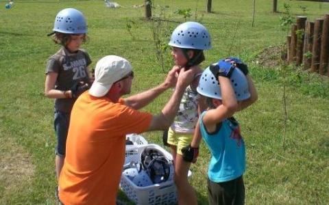 Ein Erwachsener hilft einer Gruppe von Kindern beim Anziehen von Helmen und Handschuhen auf einer Wiese, um sie auf eine Aktivität vorzubereiten.