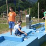 Vier Kinder spielen mit einer blauen Wasserspielstruktur auf einer Wiese mit Hügeln und Spielgeräten im Hintergrund.