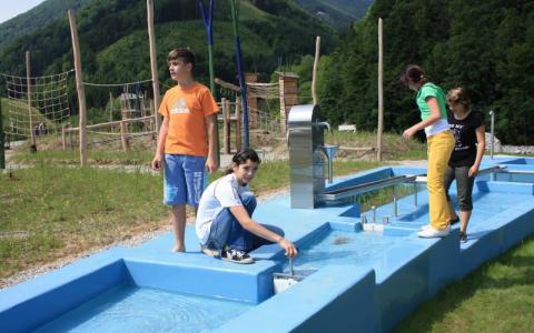 Vier Kinder spielen mit einer blauen Wasserspielstruktur auf einer Wiese mit Hügeln und Spielgeräten im Hintergrund.