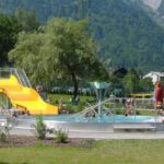 Die Menschen genießen ein öffentliches Freibad mit einer gelben Rutsche, umgeben von Gras, Bäumen und Bergen im Hintergrund.