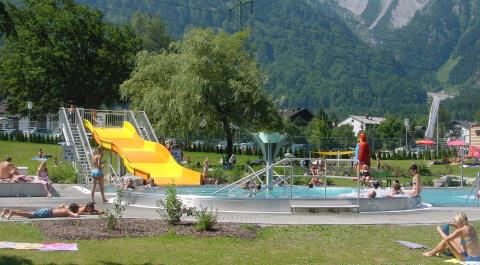 Die Menschen genießen ein öffentliches Freibad mit einer gelben Rutsche, umgeben von Gras, Bäumen und Bergen im Hintergrund.