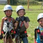 Drei Kinder mit Helmen und Klettergurten stehen draußen auf der Wiese und bereiten sich auf einen Hochseilgarten oder eine Kletteraktivität vor.