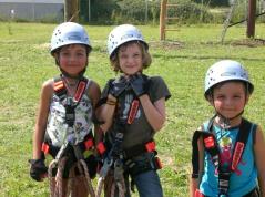 Drei Kinder mit Helmen und Klettergurten stehen draußen auf der Wiese und bereiten sich auf einen Hochseilgarten oder eine Kletteraktivität vor.