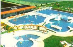 Luftaufnahme eines öffentlichen Freibadkomplexes mit mehreren Becken, einer Rutsche, einer Brücke, grünen Rasenflächen und einem nahe gelegenen Gebäude.