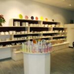 Ein Töpferatelier mit Regalen mit unbemalten Keramiken, Farbflaschen auf einer runden Auslage und einem Blumenarrangement auf dem hinteren Regal.