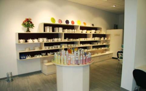 Ein Töpferatelier mit Regalen mit unbemalten Keramiken, Farbflaschen auf einer runden Auslage und einem Blumenarrangement auf dem hinteren Regal.