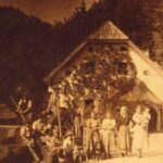 Ein sepiafarbenes Foto zeigt eine Gruppe von Menschen, die vor einem Haus mit steilem Dach, umgeben von Bäumen, posieren.