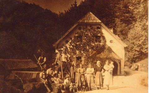 Ein sepiafarbenes Foto zeigt eine Gruppe von Menschen, die vor einem Haus mit steilem Dach, umgeben von Bäumen, posieren.