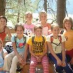 Acht Kinder sitzen und stehen gemeinsam auf einer Bank im Freien, in Freizeitkleidung und mit Rucksäcken, mit Bäumen und Sonnenlicht im Hintergrund.