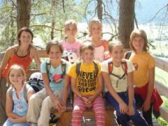 Acht Kinder sitzen und stehen gemeinsam auf einer Bank im Freien, in Freizeitkleidung und mit Rucksäcken, mit Bäumen und Sonnenlicht im Hintergrund.