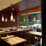 Ein modernes, leeres Café mit Holztischen und -stühlen, Hängeleuchten und großen Fenstern mit Blick auf einen Sitzbereich im Freien und Grünflächen.