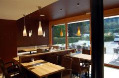 Ein modernes, leeres Café mit Holztischen und -stühlen, Hängeleuchten und großen Fenstern mit Blick auf einen Sitzbereich im Freien und Grünflächen.