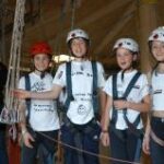 Vier Kinder mit Helmen und Klettergurten stehen in der Halle und lächeln in die Kamera, während um sie herum Seile und Ausrüstung zu sehen sind.