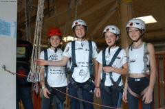 Vier Kinder mit Helmen und Klettergurten stehen in der Halle und lächeln in die Kamera, während um sie herum Seile und Ausrüstung zu sehen sind.