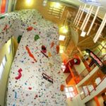 Indoor-Kletterhalle mit einer hohen, mehrfarbigen künstlichen Kletterwand, roten Geländern und einer Treppe in einem hell erleuchteten Raum.