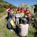 Eine Gruppe von Kindern und Erwachsenen versammelt sich im Freien auf einem grasbewachsenen Hügel und hört einem sitzenden Lehrer zu, der ein Whiteboard hält.