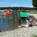 Zwei Mädchen in Badeanzügen sitzen an einem felsigen Seeufer und lächeln in die Kamera. Im Hintergrund sind Paddelboote und Menschen im Wasser und auf dem grasbewachsenen Ufer zu sehen.