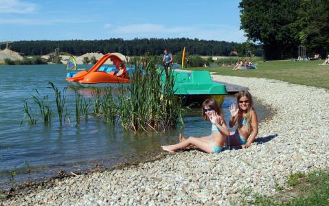 Zwei Mädchen in Badeanzügen sitzen an einem felsigen Seeufer und lächeln in die Kamera. Im Hintergrund sind Paddelboote und Menschen im Wasser und auf dem grasbewachsenen Ufer zu sehen.
