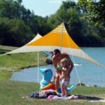 Ein Erwachsener und ein Kind stehen in der Nähe eines Sees unter einem gelb-weißen Vordach, in der Nähe liegen ein blaues aufblasbares Tier und Handtücher im Gras.