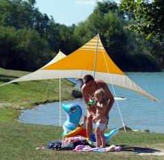Ein Erwachsener und ein Kind stehen in der Nähe eines Sees unter einem gelb-weißen Vordach, in der Nähe liegen ein blaues aufblasbares Tier und Handtücher im Gras.