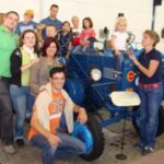 Eine Gruppe von Menschen, darunter Kinder und Erwachsene, posieren gemeinsam um einen blauen Oldtimer-Traktor in einem Innenraum. Einige Kinder sitzen auf dem Traktor, während andere in der Nähe stehen.