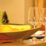 Ein Holztisch mit zwei leeren Weingläsern, einer gefalteten kastanienbraunen Serviette und einer Obstschale mit Bananen, Orangen und Grünzeug.