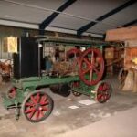 Eine alte grün-rote dampfbetriebene Landmaschine ist neben anderen antiken landwirtschaftlichen Geräten ausgestellt.