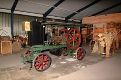 Eine alte grün-rote dampfbetriebene Landmaschine ist neben anderen antiken landwirtschaftlichen Geräten ausgestellt.