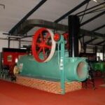 Eine große alte Dampfmaschine mit rotem Schwungrad steht in einem Innenraum auf einer gemauerten Plattform, umgeben von Industrieanlagen.