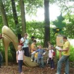 Mehrere Kinder und Erwachsene stehen neben einer großen goldenen Stöckelschuhskulptur in einem bewaldeten Außenbereich.