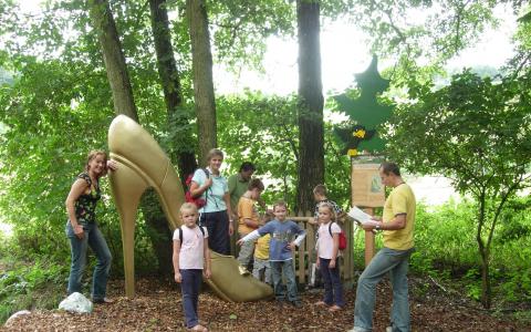 Mehrere Kinder und Erwachsene stehen neben einer großen goldenen Stöckelschuhskulptur in einem bewaldeten Außenbereich.