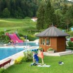 Freibadkomplex mit Menschenschwimmen, Wasserrutschen und einer kleinen Holzhütte, umgeben von Gras, Bäumen und Hügeln im Hintergrund.