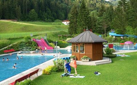 Freibadkomplex mit Menschenschwimmen, Wasserrutschen und einer kleinen Holzhütte, umgeben von Gras, Bäumen und Hügeln im Hintergrund.