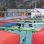 Auf einem aufblasbaren Spielfeld im Freien spielen Menschen Tischfußball, die jeweils an Schiebestangen befestigt sind, während im Hintergrund Berge und Gebäude zu sehen sind.