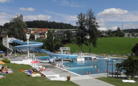 Öffentliches Freibad mit Wasserrutschen, Menschen, die sich auf der Wiese sonnen, und Schwimmer im Becken; Wohngebäude und Bäume im Hintergrund unter einem teilweise bewölkten Himmel.