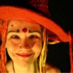 Lächelnde Person, die einen großen roten Hut mit Garn- oder Stofffäden und bunter Gesichtsbemalung trägt, darunter ein roter Punkt auf der Stirn und blaue Akzente über den Augen.