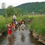 Mehrere Kinder und ein Erwachsener waten durch einen seichten Bach in einer ländlichen Gegend mit grünen Feldern, Bäumen und Hügeln im Hintergrund.