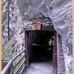 Hölzerner Eingang zu einem engen Tunnel, der in einen Felsen gebaut ist, mit einem Holzgeländer auf der linken Seite und einem kleinen Schild über dem Eingang.