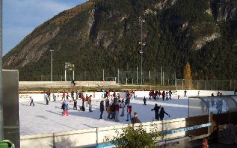 Menschen laufen Schlittschuh auf einer von einem Zaun umgebenen Eisbahn im Freien, im Hintergrund sind Berge und Bäume zu sehen.