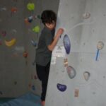 Ein Junge klettert an einer Indoor-Boulderwand, hält sich an bunten Griffen fest und steht auf einer blauen Matte.