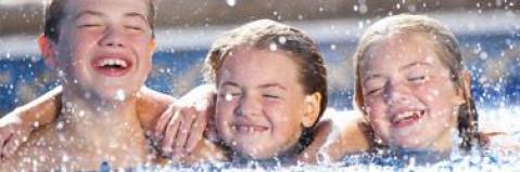 Drei Kinder mit nassen Haaren lächeln und halten sich gegenseitig in einem Schwimmbad, während das Wasser um sie herum spritzt.