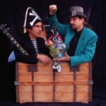 Zwei Männer in Kostümen, einer als Pirat mit Banjo, der andere mit blaugrüner Jacke und grauem Hut, stehen hinter einer offenen Holztruhe mit einem Fischglas und einer Taschenuhr.